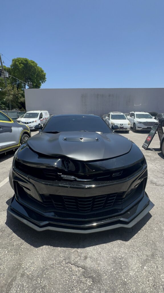 Luxury sports car with front-end collision damage, missing bumper and grille, parked outside Primo Paint & Body